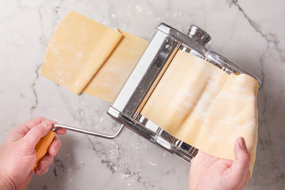 Fresh pasta sheets being rolled through a pasta machine to create thin sheets for lobster ravioli. The pasta roller is clamped to the counter as dough passes through the rollers to achieve the proper thickness for filled pasta.