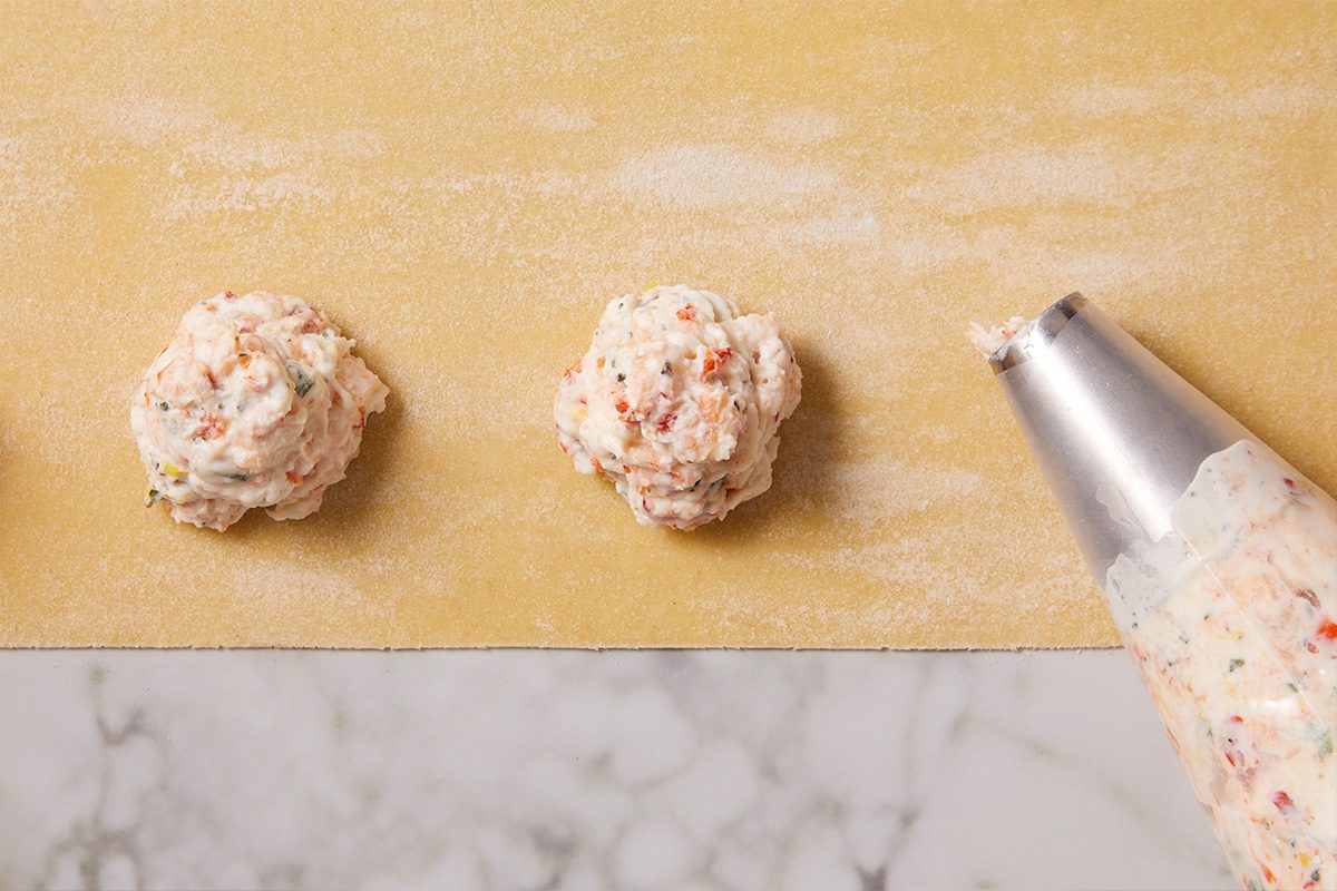 Lobster filling being piped onto a sheet of pasta dough in evenly spaced portions using a piping bag, preparing the filling placement before sealing and cutting ravioli.
