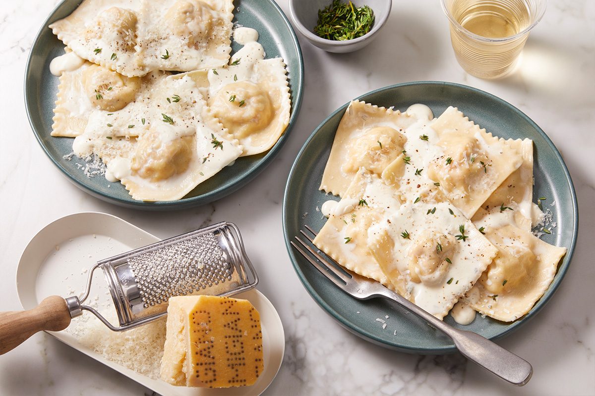 Two plates of lobster ravioli served with creamy sauce and fresh herbs, styled for entertaining with grated cheese and wine in the background.