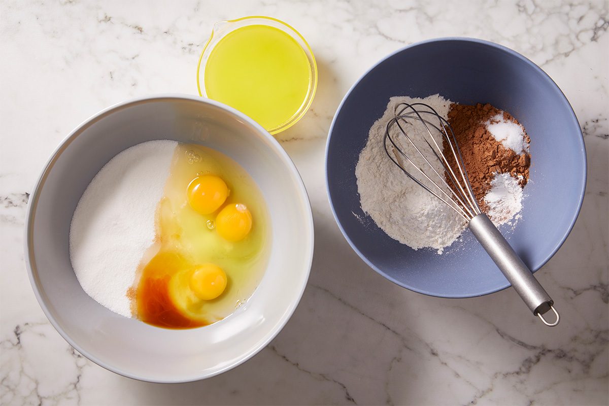 Overhead view of sugar, eggs and vanilla extract in a mixing bowl alongside melted butter and a separate bowl of dry ingredients for Lunch Lady Brownies. The ingredients are staged and ready to be combined for this classic chocolate sheet pan dessert.
