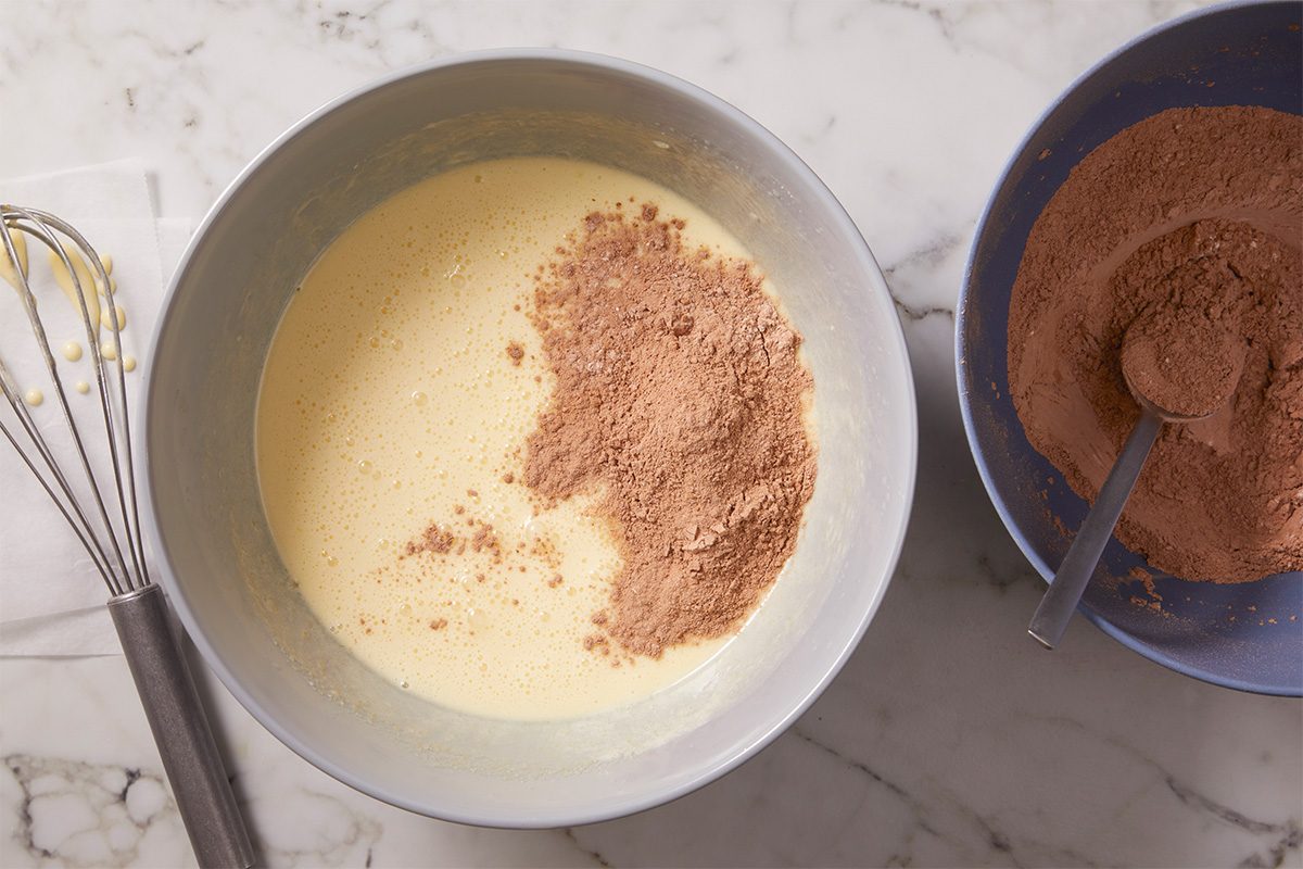 Overhead image of brownie batter being mixed as dry ingredients—flour, baking cocoa, baking powder and salt—are added to the wet mixture. The batter begins to thicken into a smooth chocolate base.