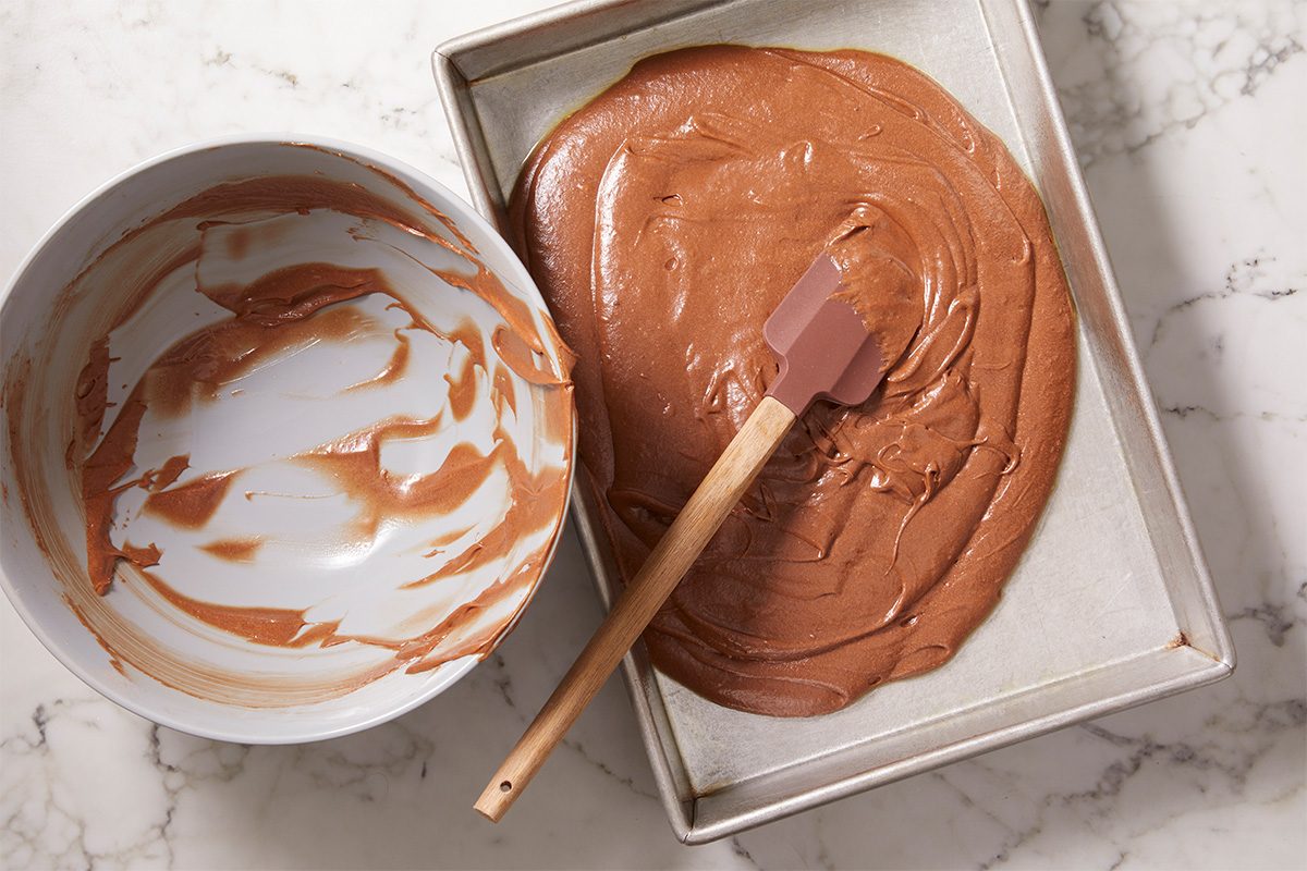 Overhead view of thick chocolate brownie batter spread evenly into a rectangular baking pan with a spatula, ready for baking. The smooth surface ensures even cooking for fudgy, tender brownies.