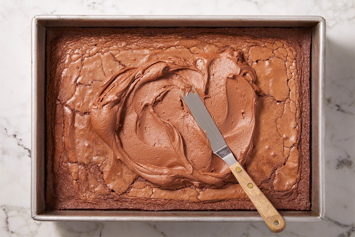Vertical overhead image of baked Lunch Lady Brownies in a rectangular pan generously topped with smooth chocolate frosting. A small offset spatula rests on the surface, highlighting the rich, glossy finish.
