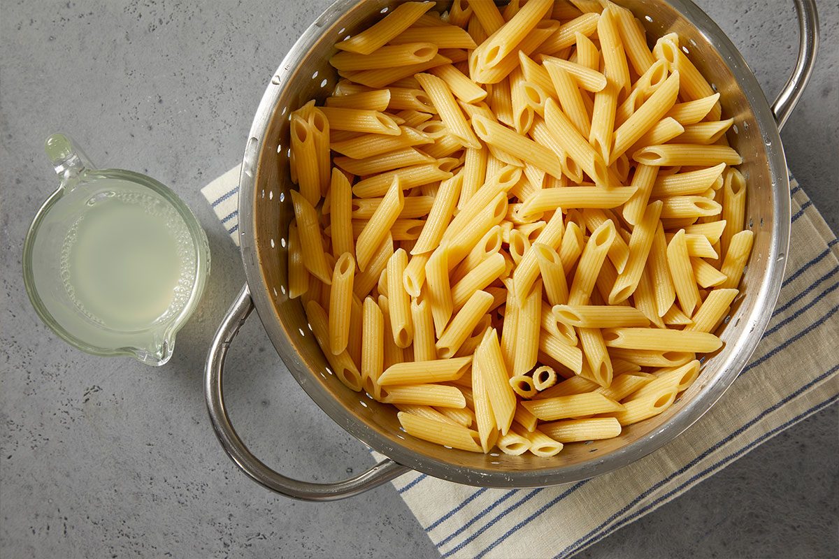 Overhead horizontal image of a colander filled with cooked penne pasta, with a small glass jug of pasta water off to the side. The colander sits on a striped kitchen towel against a grey surface.