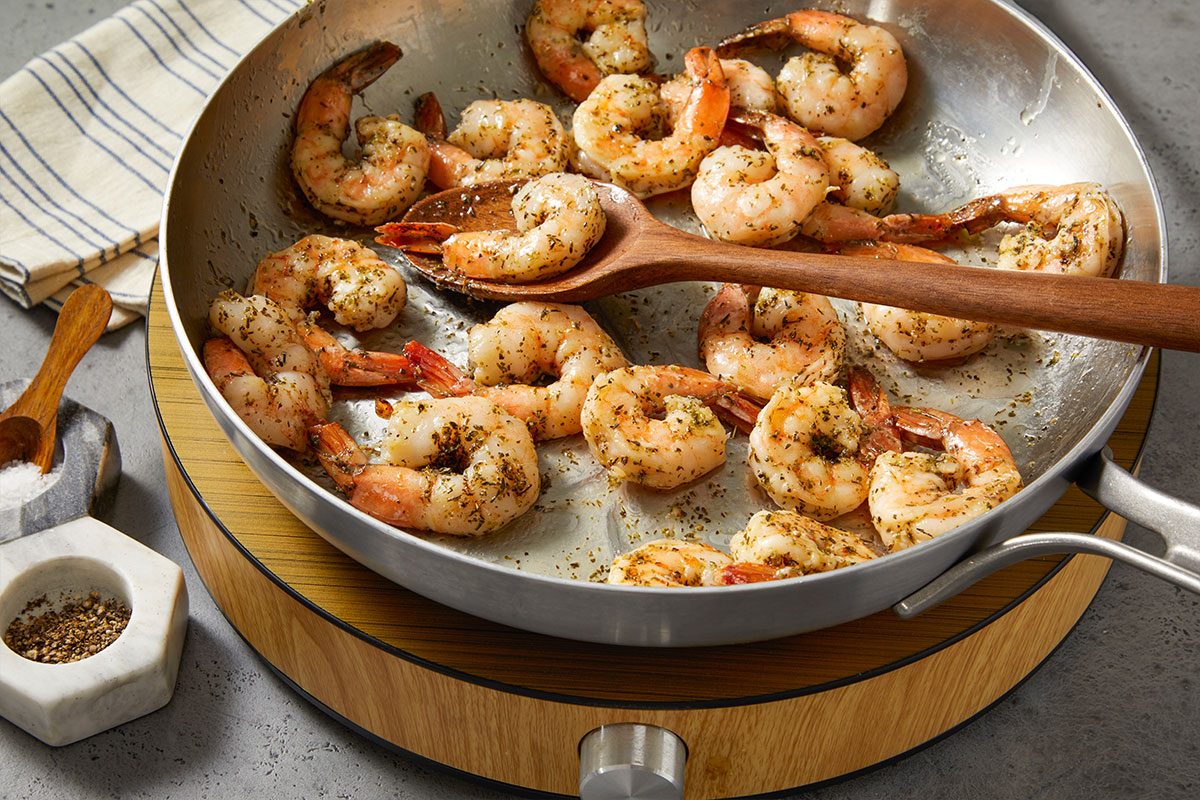 Eye Level horizontal image of cooked shrimp being tossed in a silver skillet with a wooden spoon. A bowl of pepper and a bowl of salt are also visible.