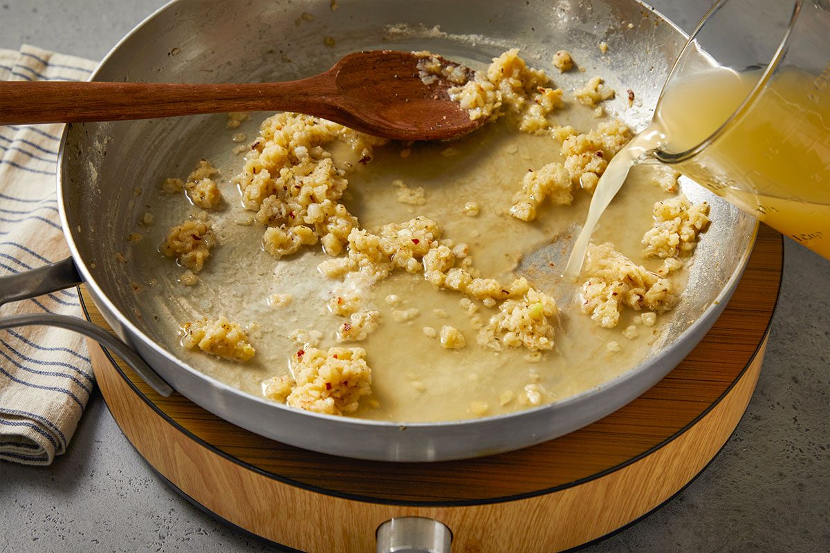 Eye Level horizontal image of a cooking pan with browned bits of food inside and a wooden spoon. Liquid is being poured into the pan from a measuring cup. The pan rests on a wooden trivet.