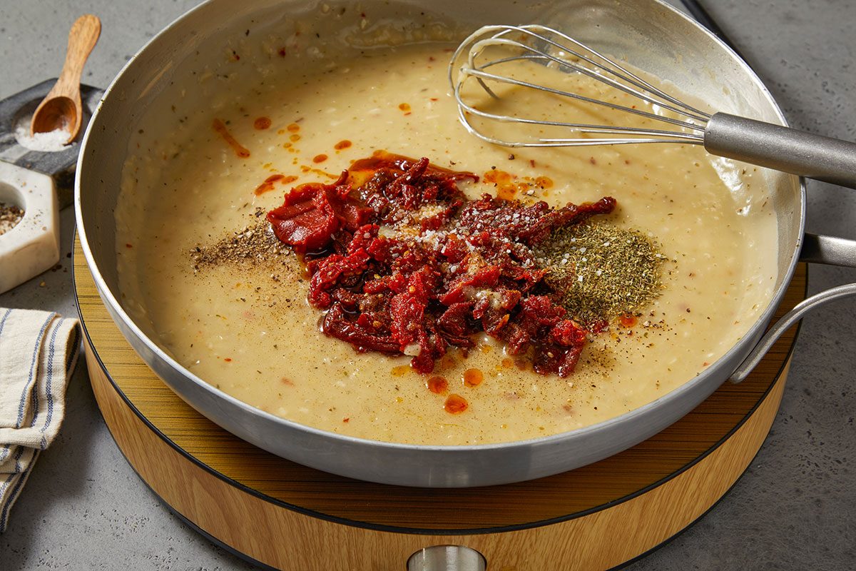 Overhead horizontal image of the addition of ingredients to a frying pan with sauce. The visible ingredients being added are sun-dried tomatoes, salt, oregano, and pepper. A whisk is in the upper right corner of the pan.