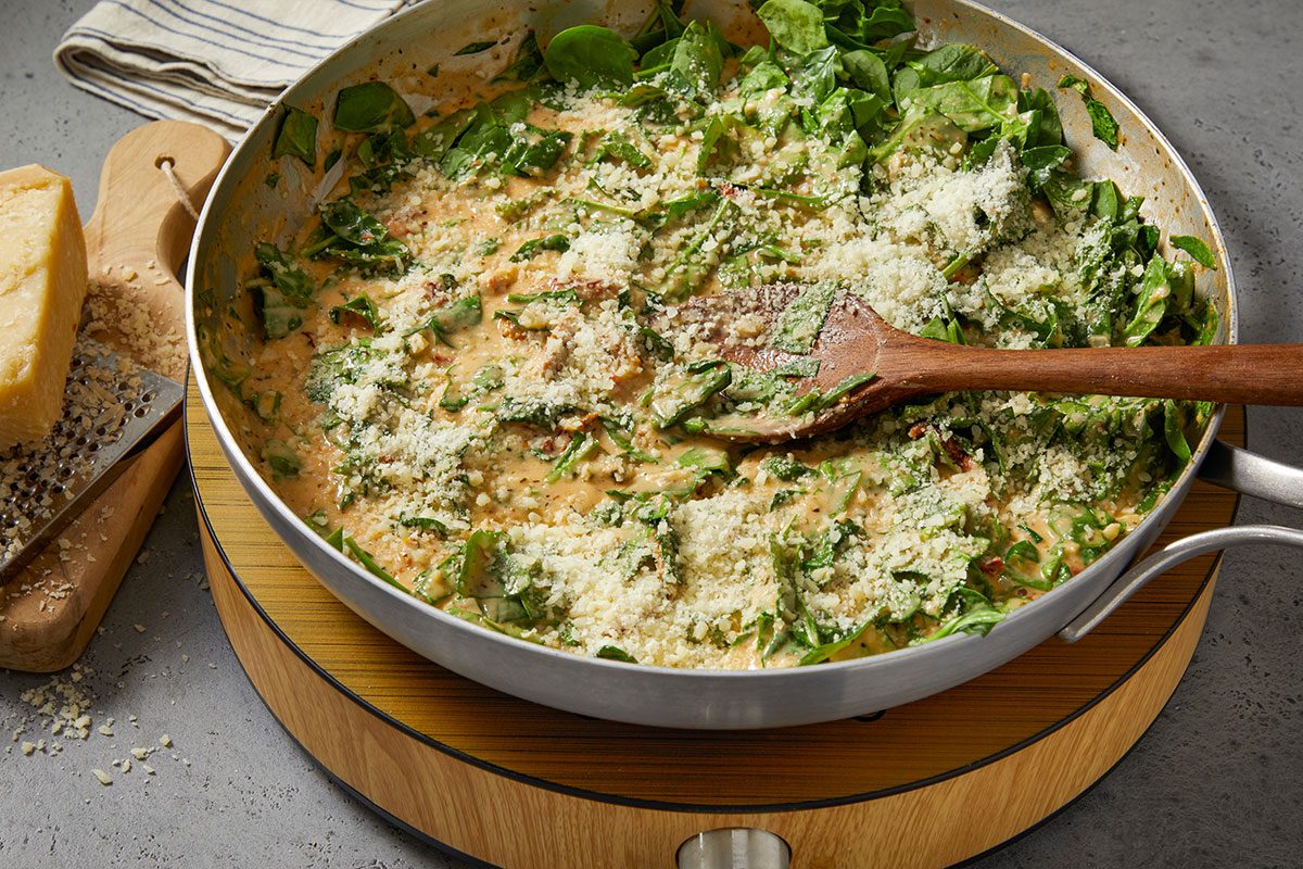 Overhead horizontal image of spinach in a skillet. A wooden spoon rests on the spinach, which is covered in shredded parmesan cheese and a cream sauce.
