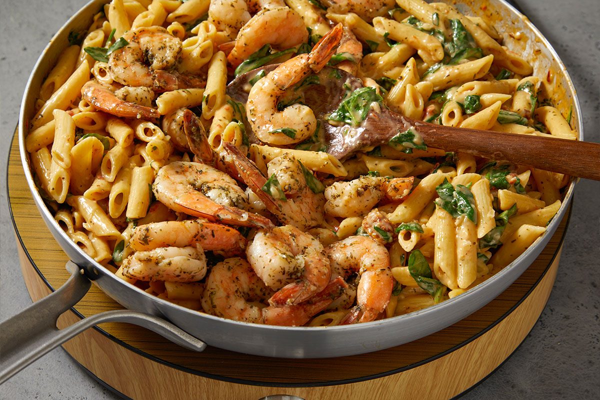 Eye Level horizontal image of shrimp pasta being stirred in a pan. The visible ingredients are penne pasta, sauteed shrimp, spinach, and a creamy sauce.