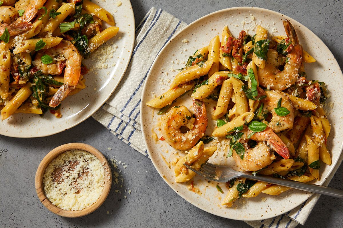 Overhead horizontal image of two plates of pasta with shrimp, basil, and spinach. A fork is piercing the pasta on one plate, and there is a small wooden bowl of cheese on the side.