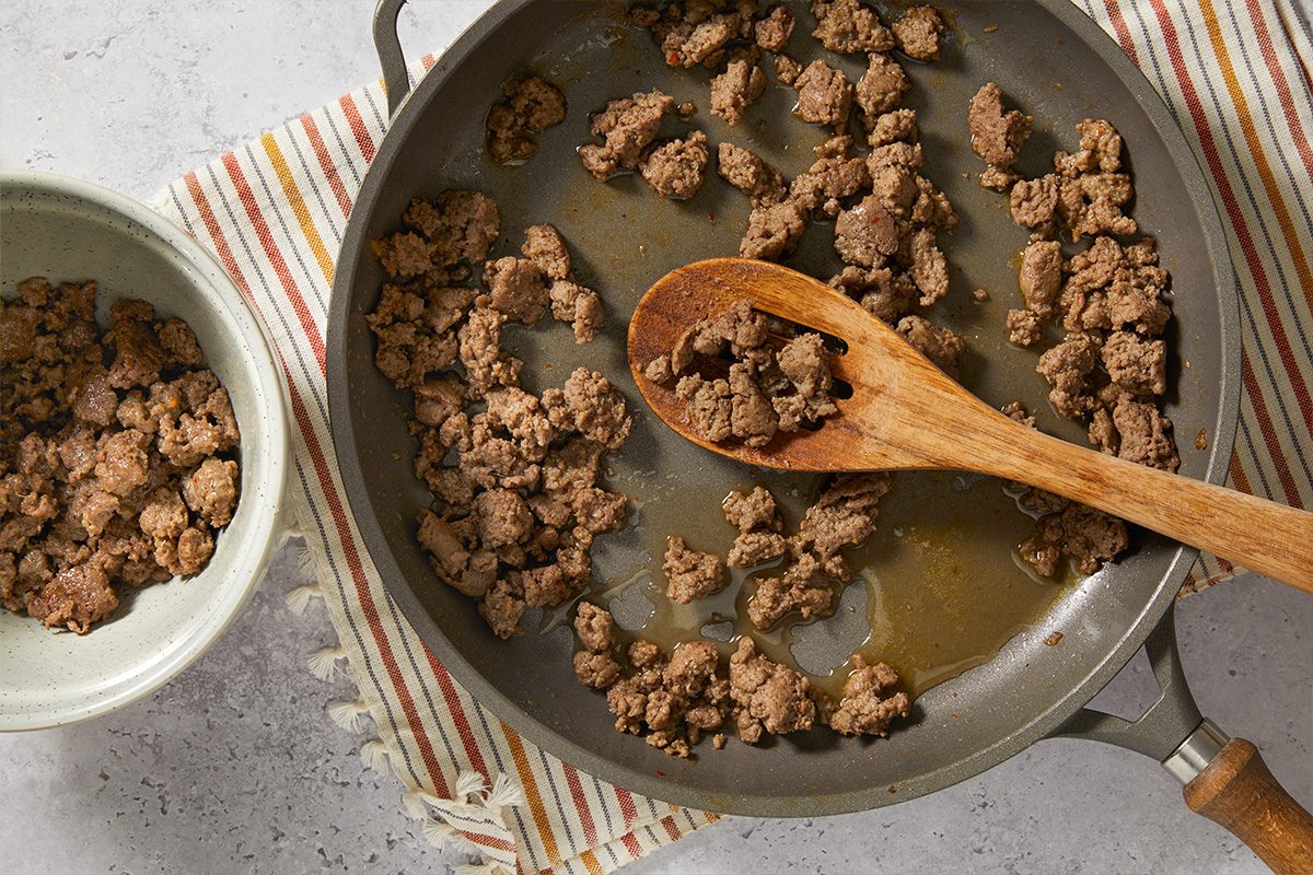 Overhead cooking shot of ground pork browning in a skillet with a wooden spoon. The crumbled pork is cooked as part of the savory filling for the McGriddle breakfast casserole.