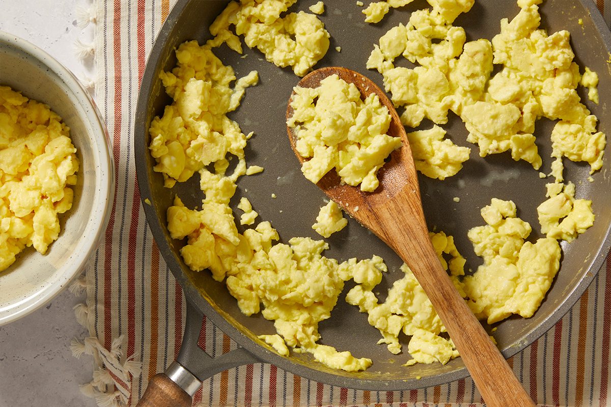 Overhead image of scrambled eggs cooking in a skillet with a wooden spoon. The fluffy eggs are prepared as a layer for the savory McGriddle breakfast casserole.