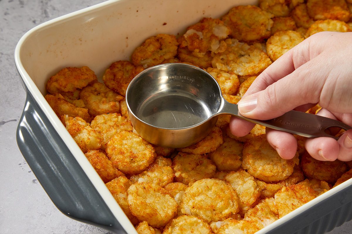 Overhead image showing a measuring cup pouring liquid mixture over a layer of tater tots in a casserole dish, beginning the assembly of the McGriddle-style breakfast casserole.