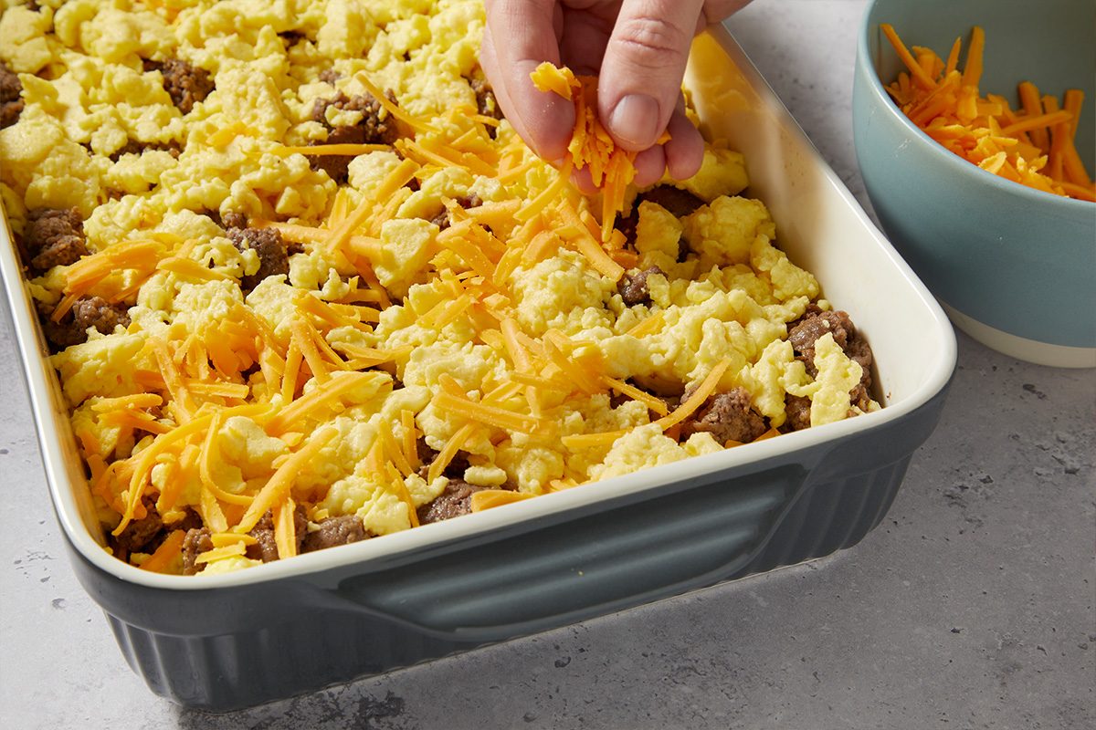 Overhead image of shredded cheese being sprinkled over the casserole ingredients in a baking dish. The cheese topping completes the layers before baking the McGriddle breakfast casserole.