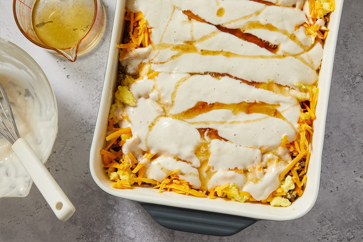 Overhead image of the fully assembled breakfast casserole topped with creamy sauce and syrup drizzle before baking. The layered ingredients are visible in the casserole dish.