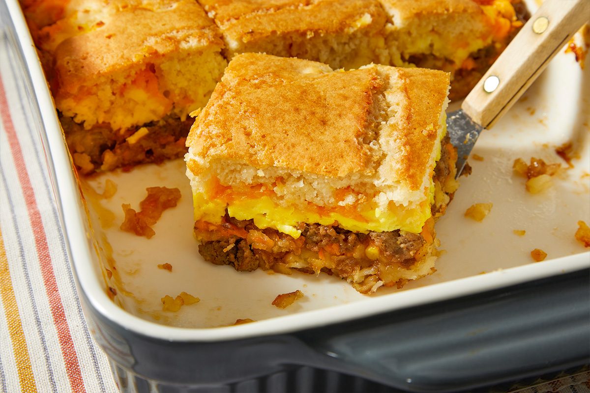 Close-up overhead view of a slice of McGriddle casserole lifted from the baking dish, showing layers of sausage, eggs and crispy potato base.