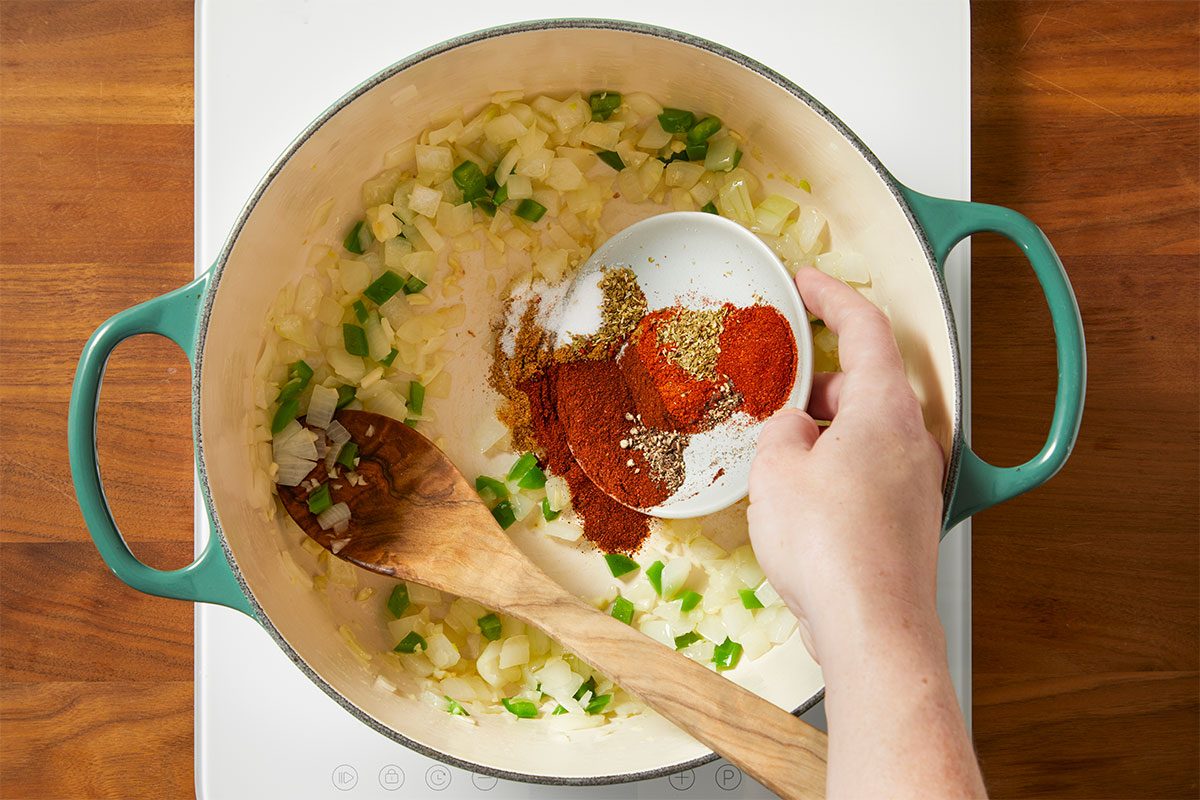 Overhead horizontal image of step 82 of 116 in a series. A hand is seen pouring a variety of spices from a small dish into a pot filled with sauteed onions and green bell peppers.