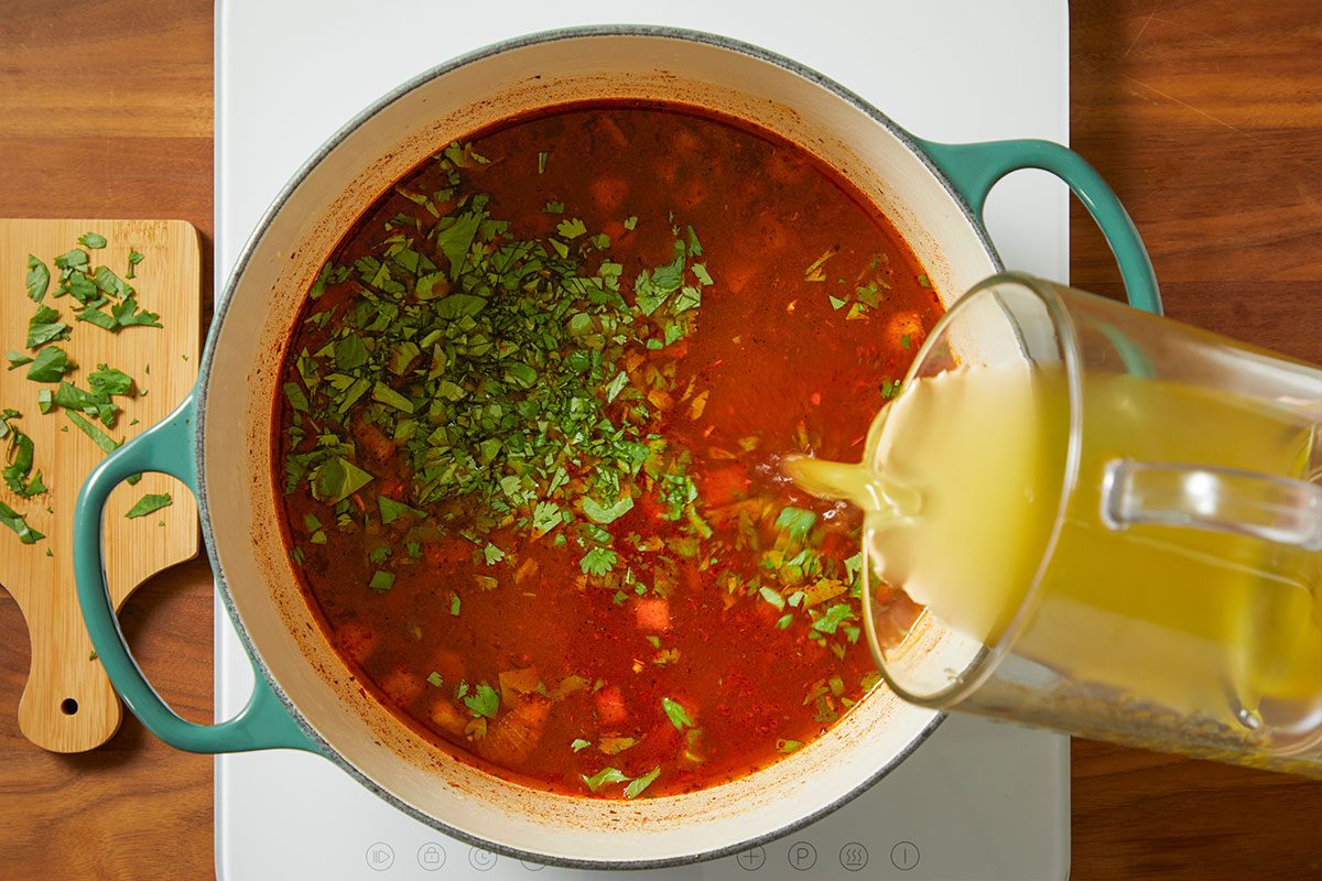 Overhead horizontal image of a pot of soup being prepared. Someone is pouring broth into the pot of soup which contains cilantro and tomatoes.