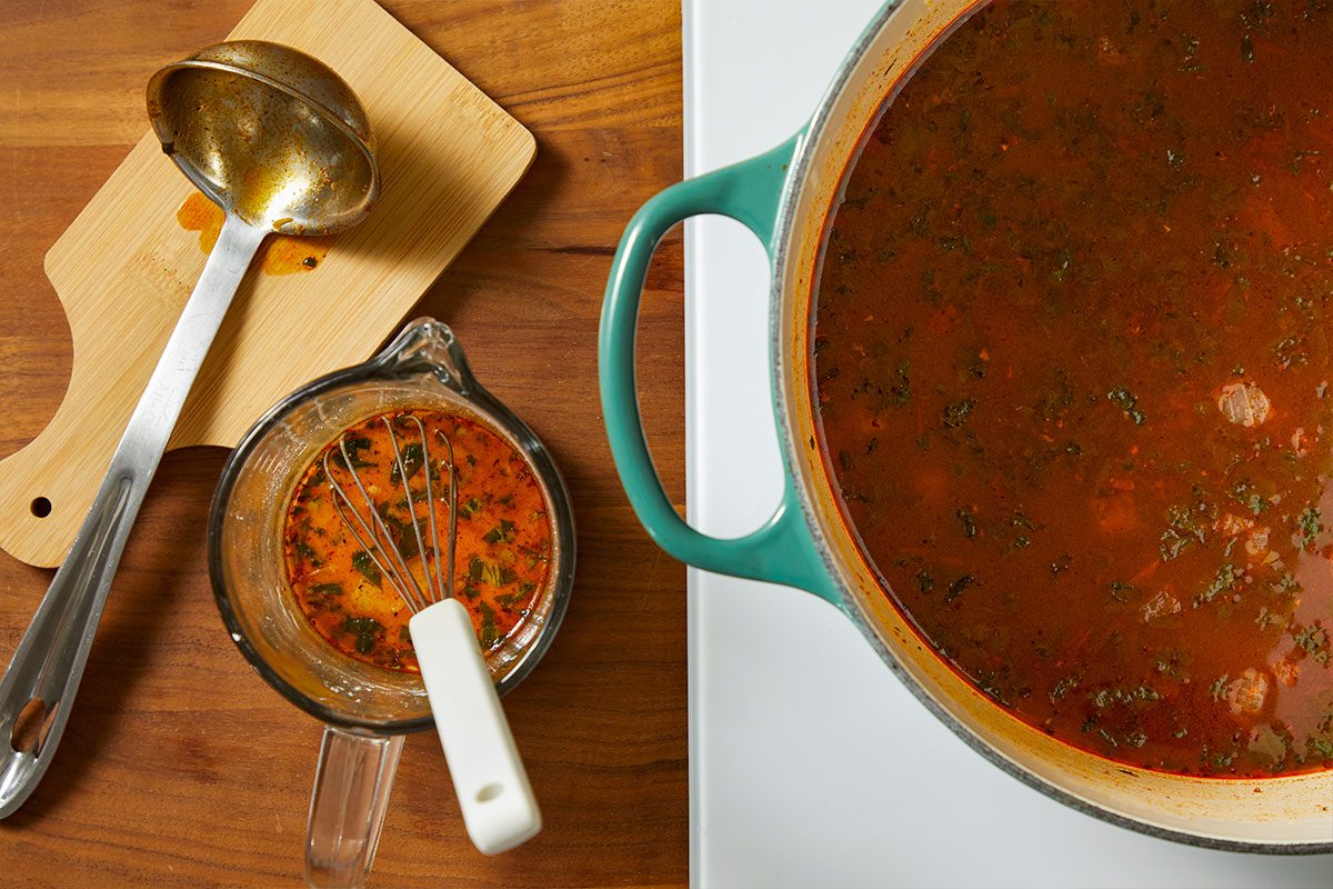 Overhead horizontal image of the preparation of soup. A measuring cup of seasoning liquid is seen next to a pot of soup and a ladle. The liquid is being whisked in a measuring cup.