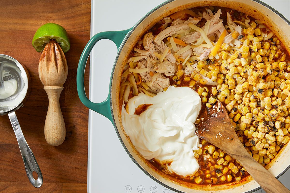 Overhead horizontal image of a pot of food with a wooden spoon, topped with sour cream, shredded cheese, corn and shredded chicken. A metal measuring cup of sour cream and lime juicer are on a wooden board next to the pot.