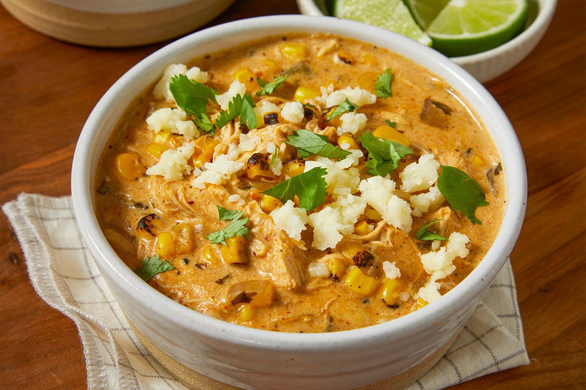 Eye level horizontal image of chicken tortilla soup in a white bowl. The soup contains grilled corn, shredded chicken, cilantro, and white cheese crumbles. Limes sit in a bowl next to the soup.