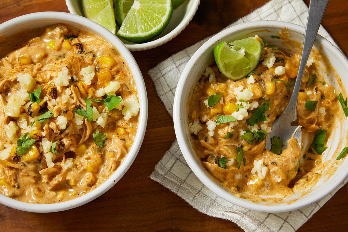 Overhead horizontal image of two bowls filled with creamy chicken and corn. The bowls are garnished with fresh cilantro, crumbled cheese, and a lime wedge with a spoon visible in one bowl.
