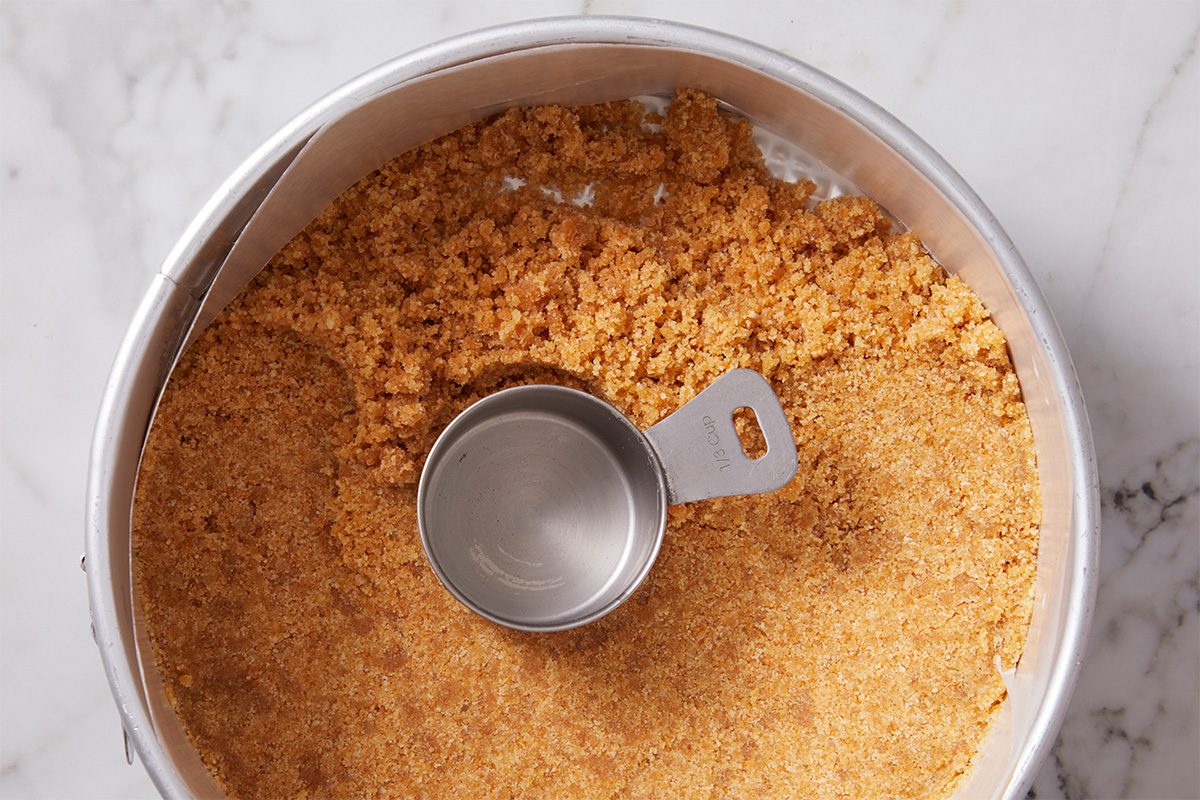 Overhead horizontal image of Alex Bala's No-Bake Banana Pudding Cheesecake. A metal baking pan is filled with vanilla wafer crust; a measuring cup sits on top of the crust.