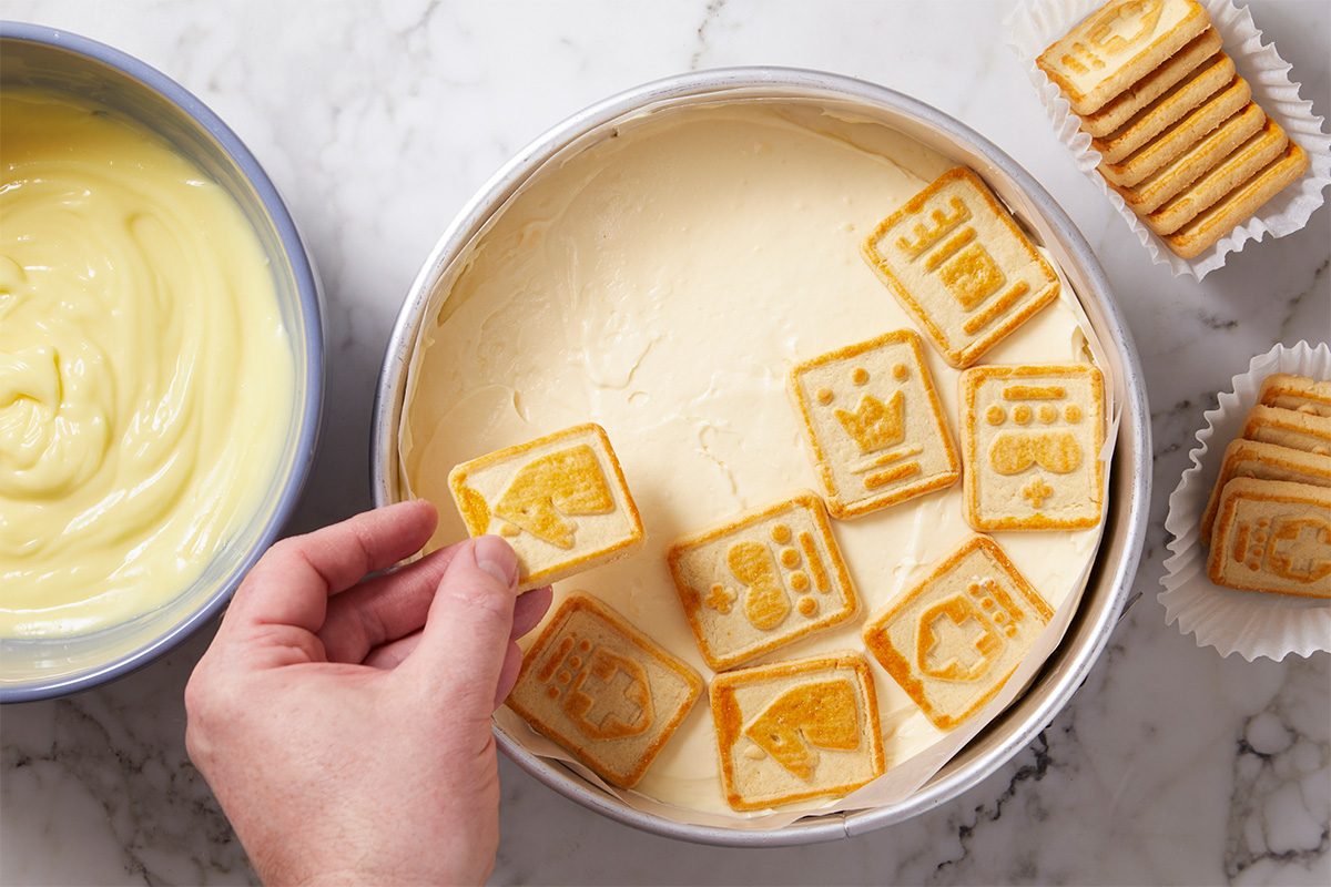 Overhead horizontal image of Alex Bala's No-Bake Banana Pudding Cheesecake, step by step. A hand is placing Chessmen cookies atop a layer of cheesecake mixture in a baking pan; more cookies are visible to the right, and pudding topping is in a bowl at the side.