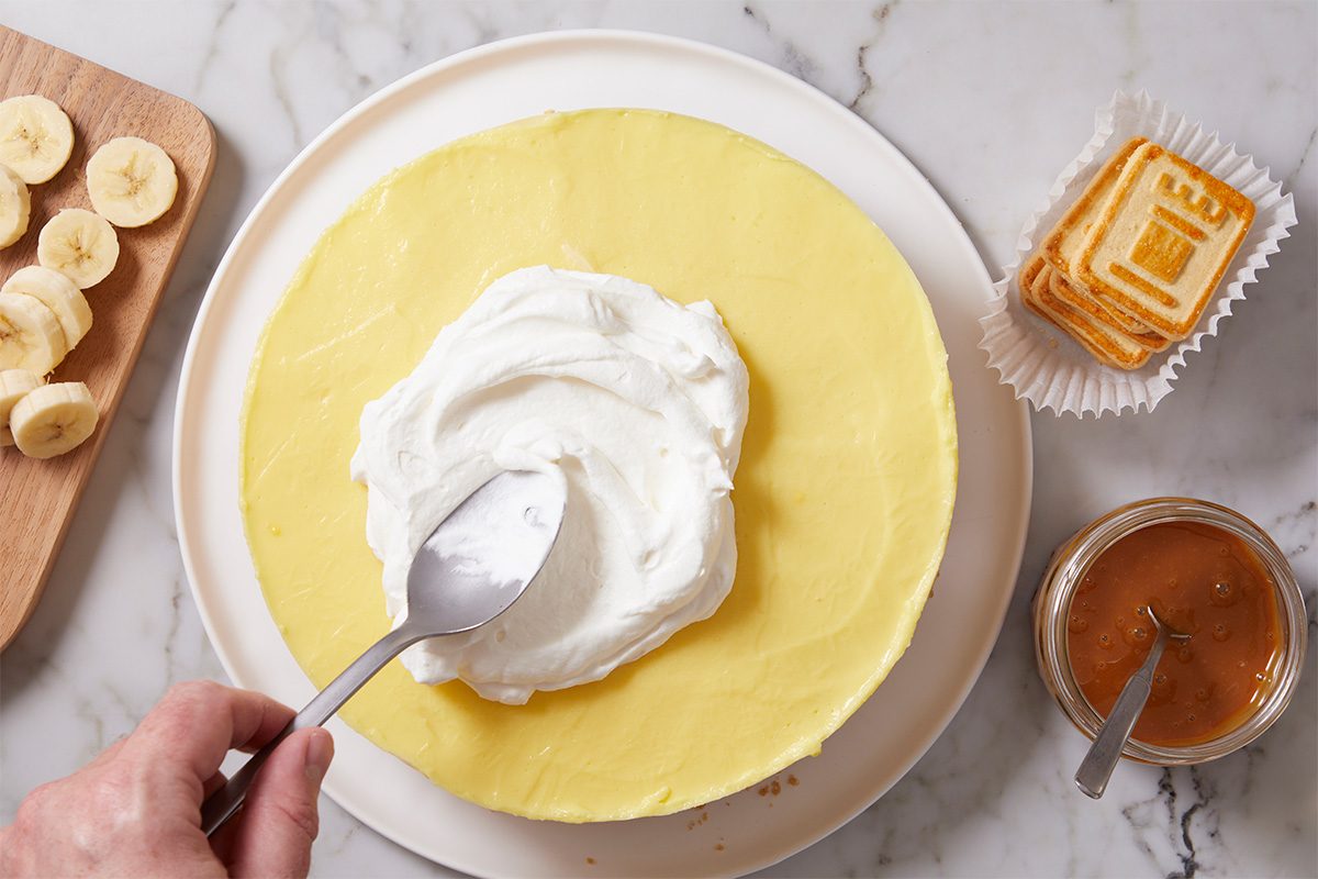 Overhead horizontal image of Alex Bala's No-Bake Banana Pudding Cheesecake. A hand uses a spoon to spread whipped topping on the pudding filling, with sliced bananas, Chessmen cookies, and caramel sauce displayed nearby.