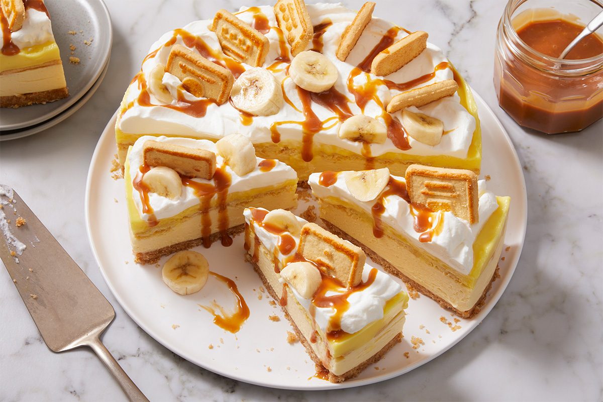 Overhead horizontal image of Alex Bala's No-Bake Banana Pudding Cheesecake, sliced and arranged on a white plate. The cheesecake is topped with whipped cream, banana slices, Chessmen cookies, and caramel sauce.