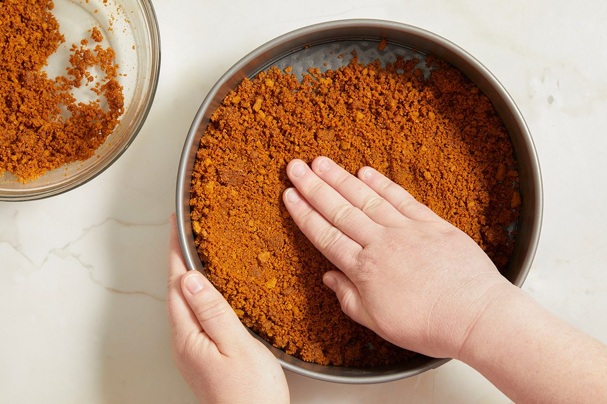 A person presses a crumbly brown graham cracker crust mixture into the bottom of a round cake pan, preparing the base for a dessert. A bowl with more crumbs sits nearby on a white surface.
