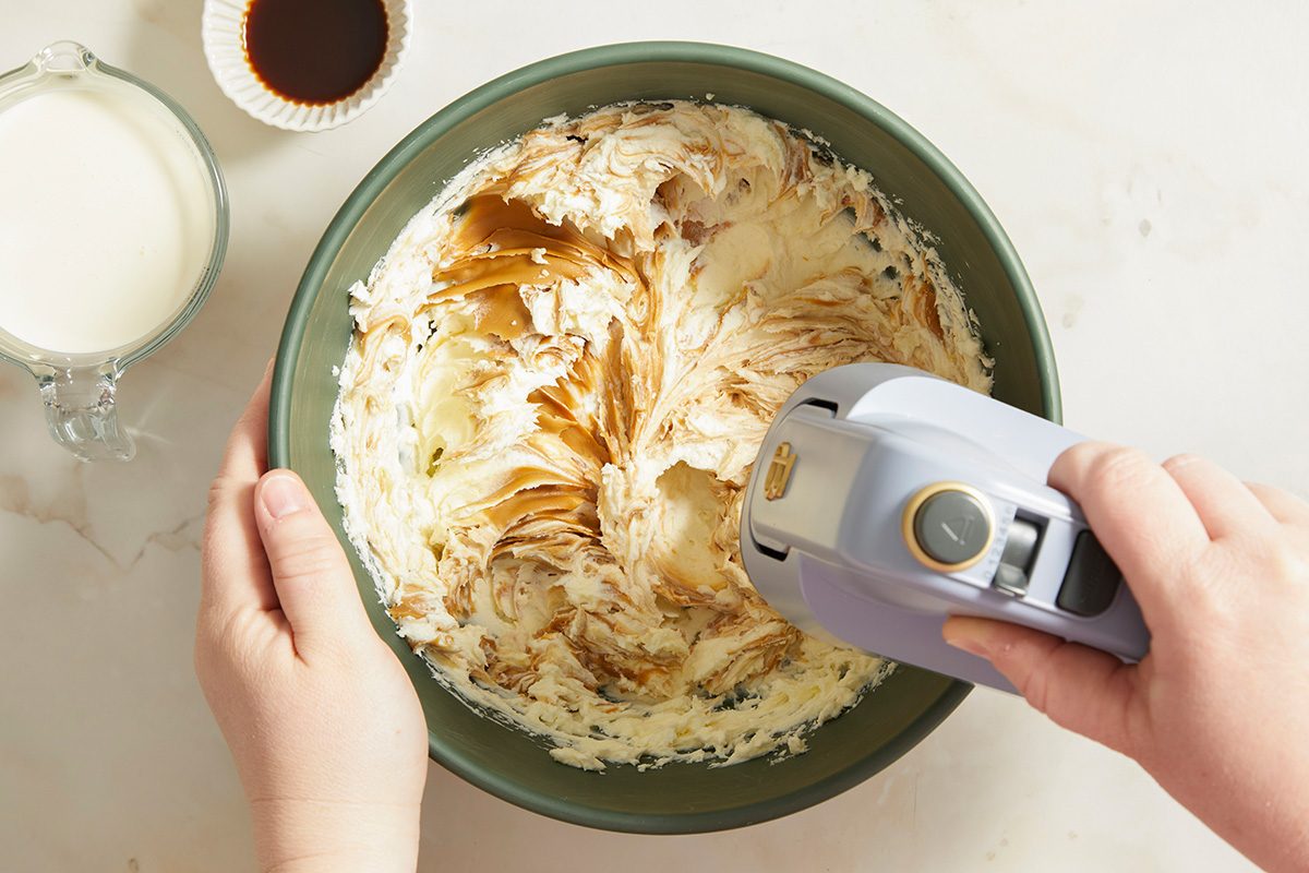 A person uses a hand mixer to blend ingredients, including cream, in a green mixing bowl. A small bowl of vanilla extract and a measuring cup of milk are nearby on a white countertop.