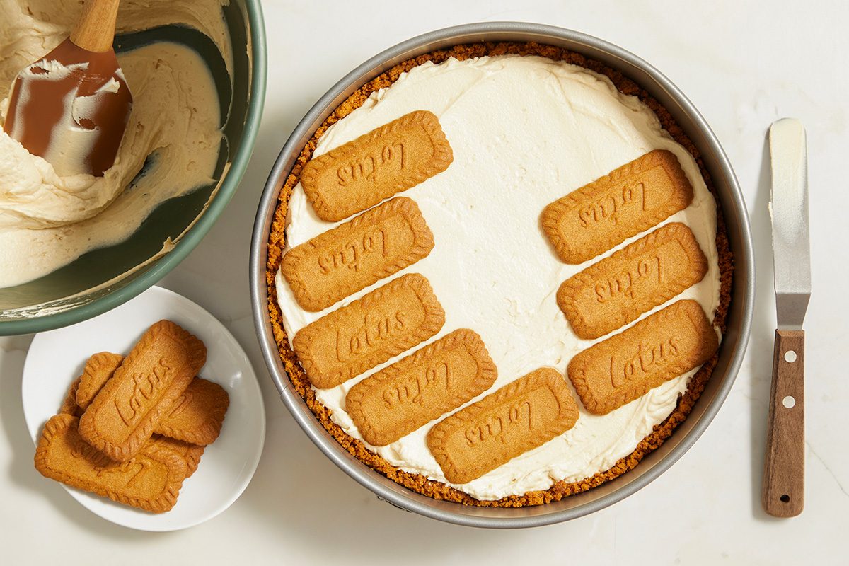 A round cheesecake topped with Lotus Biscoff cookies, with a bowl of batter, a spatula, and extra cookies on the side. A knife rests next to the cheesecake on a white surface.