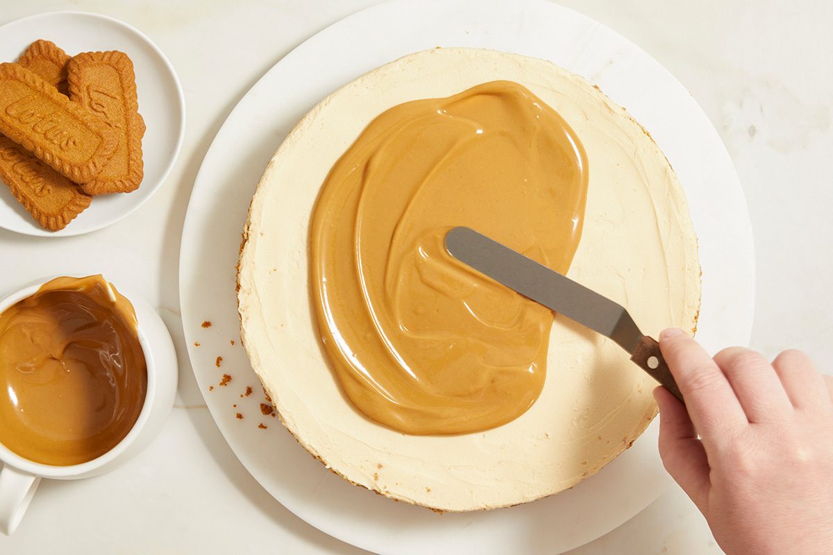 A hand uses an offset spatula to spread caramel or cookie butter on top of a round cheesecake. Nearby are a cup of the spread and a plate with cookies.