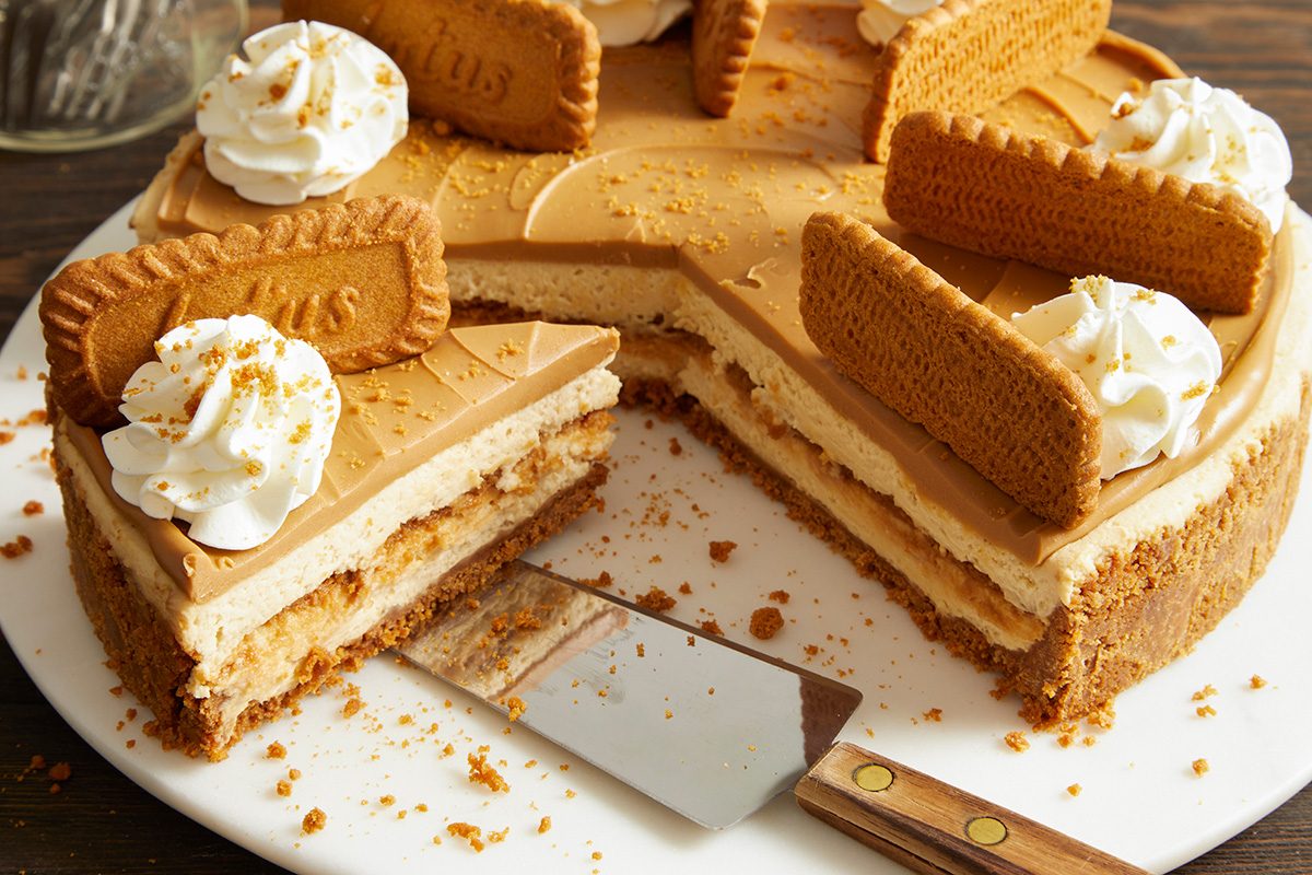 A cheesecake topped with whipped cream and rectangular cookies, with several slices cut and served. A cake server rests on the plate, and crumbs are scattered around.