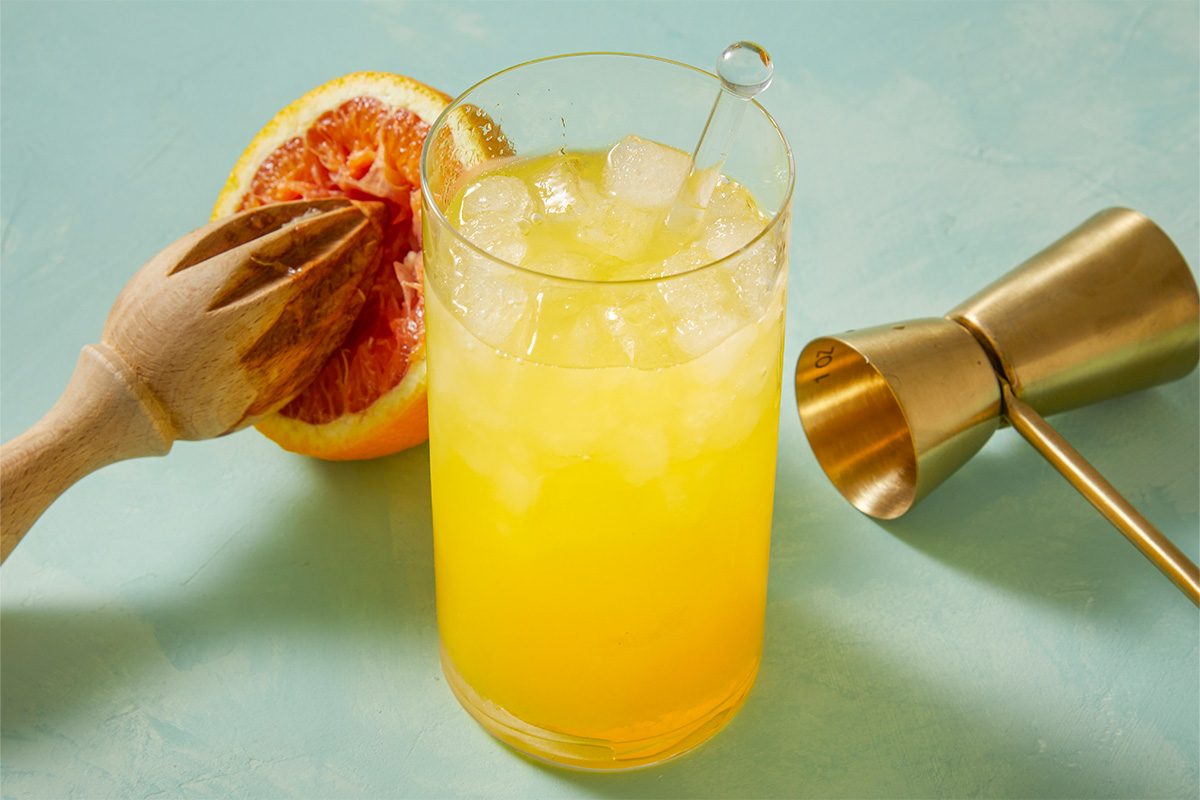 Eye Level horizontal image of a glass with a yellow liquid and ice, next to a halved orange with a wooden reamer. A golden jigger sits next to the glass.