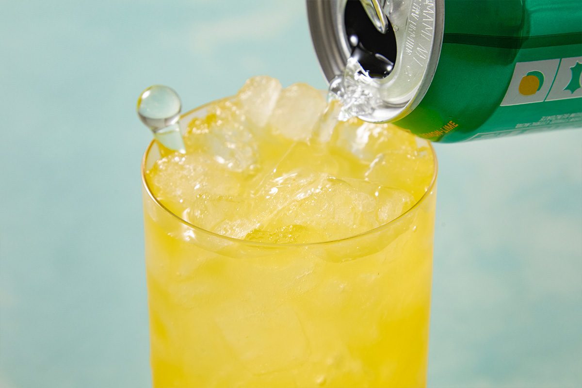Eye level horizontal image of a green lemon-lime soda can being poured into a glass filled with ice. A glass swizzle stick rests in the filled glass.