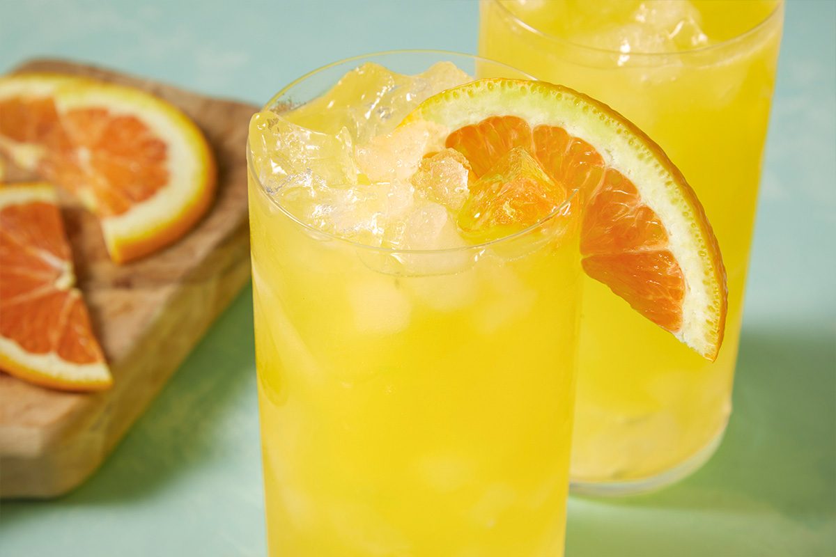 Eye Level horizontal image of a glass filled with a yellow drink and ice. The glass is garnished with an orange slice. Another glass and orange slices on a cutting board are in the background.