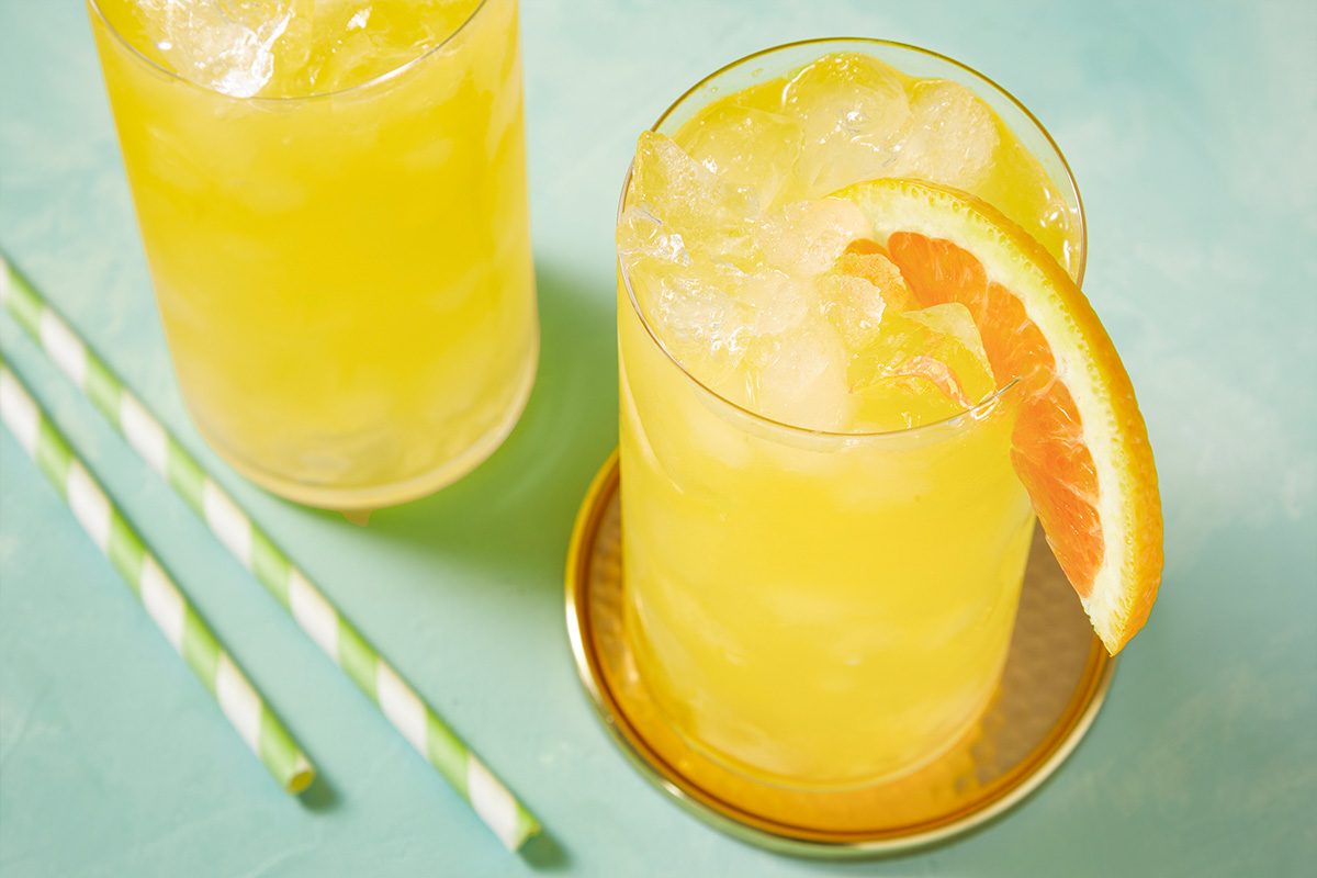 Eye Level horizontal image of two cocktails filled with ice and a yellow liquid, one garnished with an orange slice. The cocktails are served with green and white striped paper straws, placed on a light blue surface.