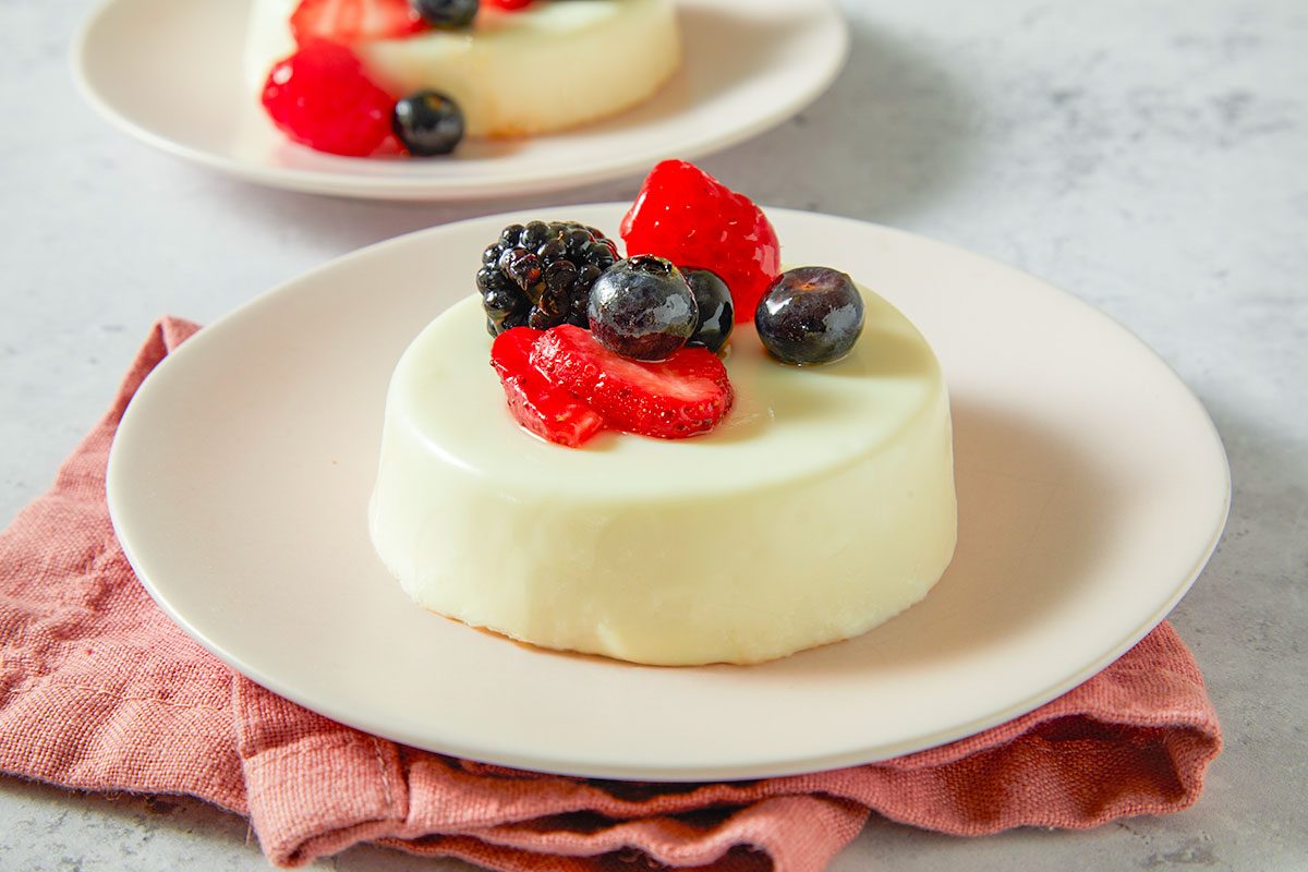 A horizontal image shows a single panna cotta topped with fresh berries on a plate.