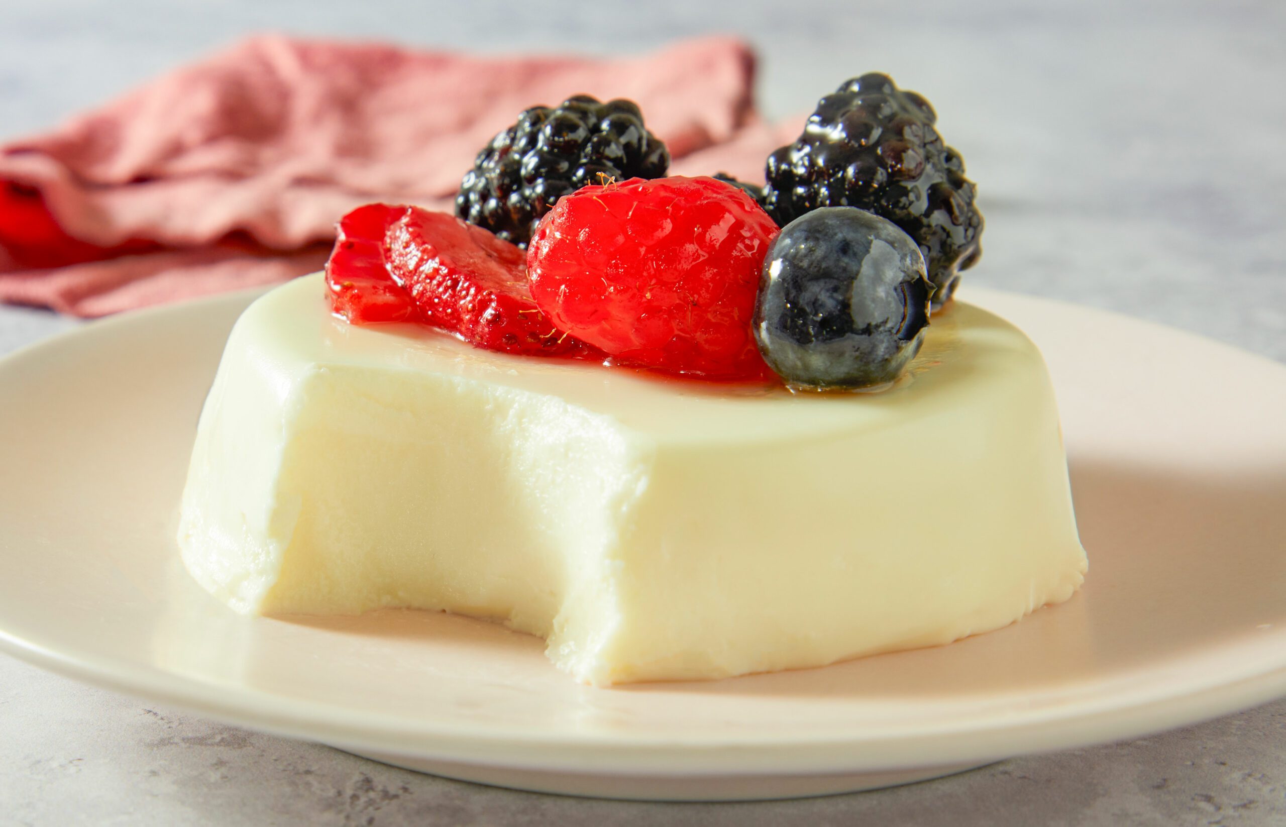 A horizontal close-up image shows panna cotta with a bite taken out, revealing its smooth, creamy texture and berry topping.