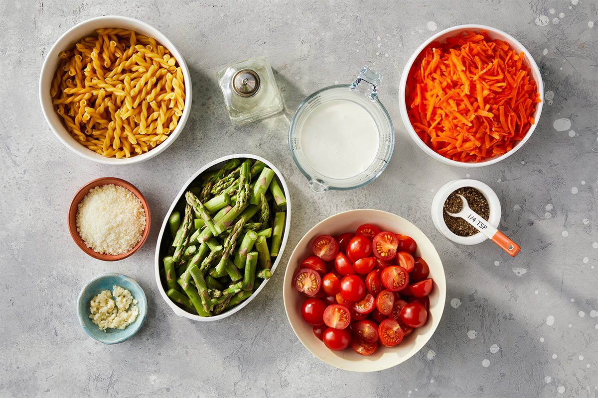 Measured ingredients including pasta, asparagus, shredded carrots, cherry tomatoes, garlic, cream, and Parmesan are arranged overhead for the creamy pasta primavera recipe.