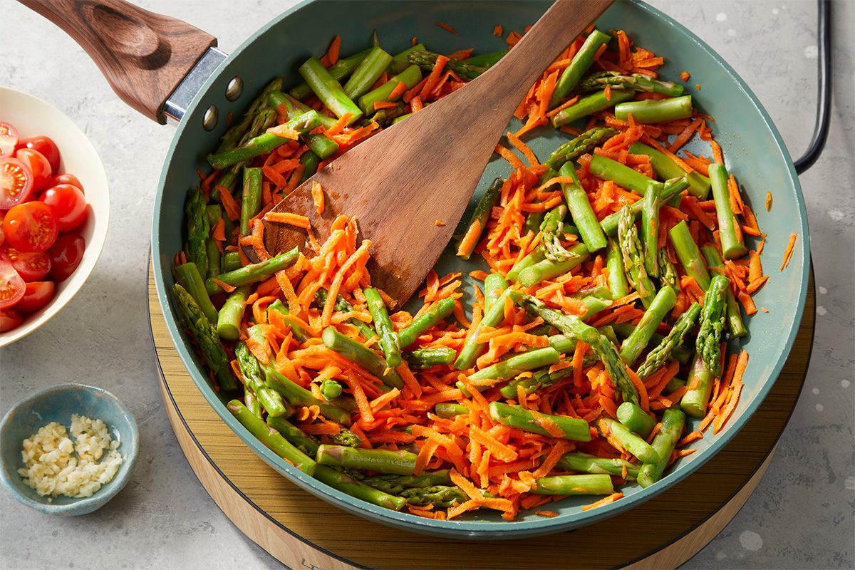 Asparagus and shredded carrots are sautéed in a skillet with oil while being stirred with a wooden spatula.