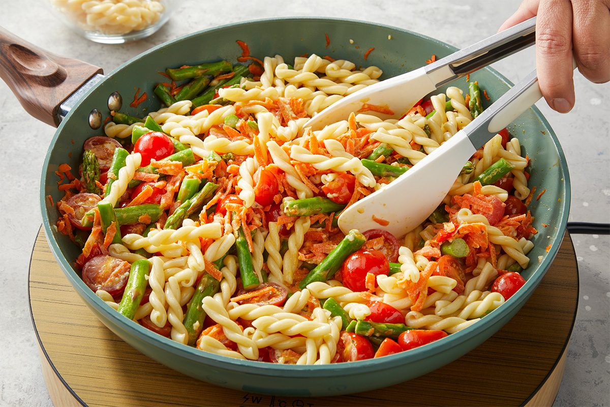 Cooked pasta is tossed with the creamy vegetable mixture in a skillet using a spatula.