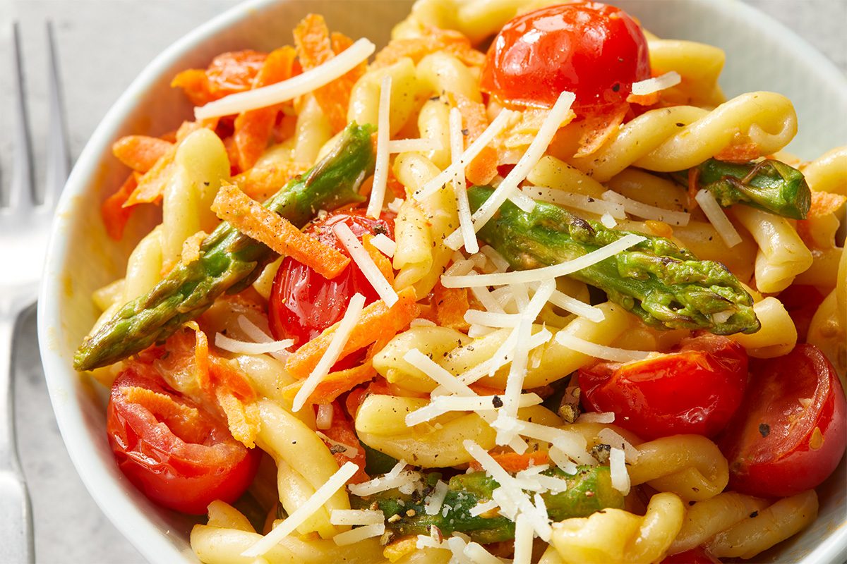 Close-up of creamy pasta primavera highlighting glossy sauce, asparagus, carrots, and cherry tomatoes with Parmesan.