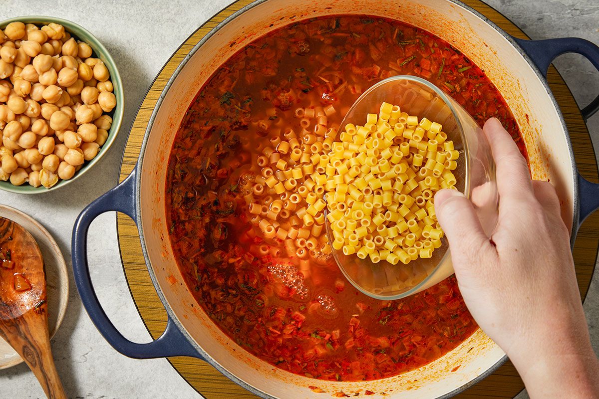Overhead horizontal image of ditalini pasta being added to simmering tomato broth with chickpeas.