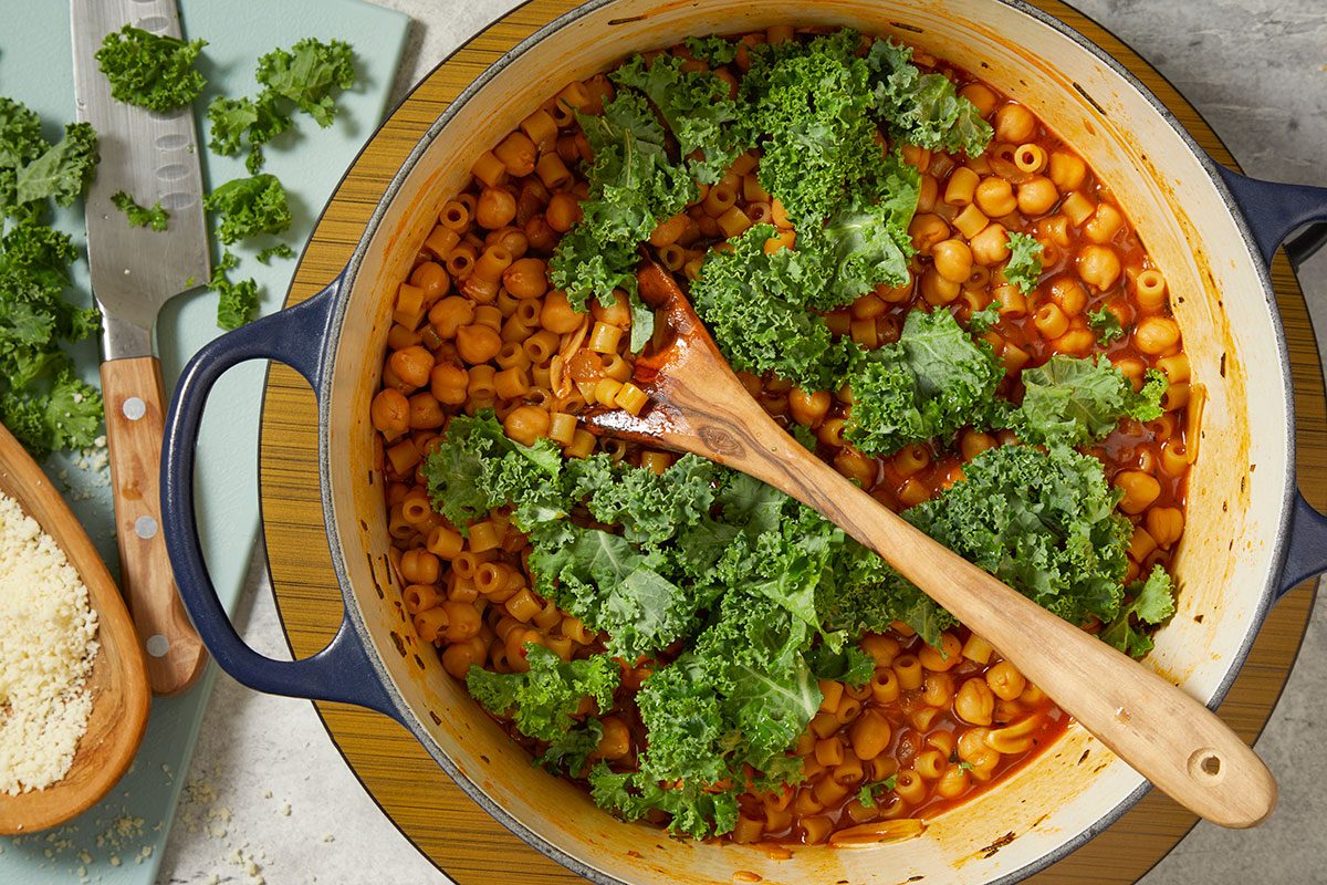 Overhead horizontal image of kale being added to pasta e ceci as it finishes cooking in a Dutch oven.