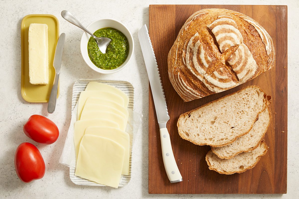 A loaf of artisan bread, two slices cut, sits on a wooden board with a bread knife. Nearby are Provolone cheese slices, two tomatoes, a stick of butter, and a bowl of green pesto with a spoon.