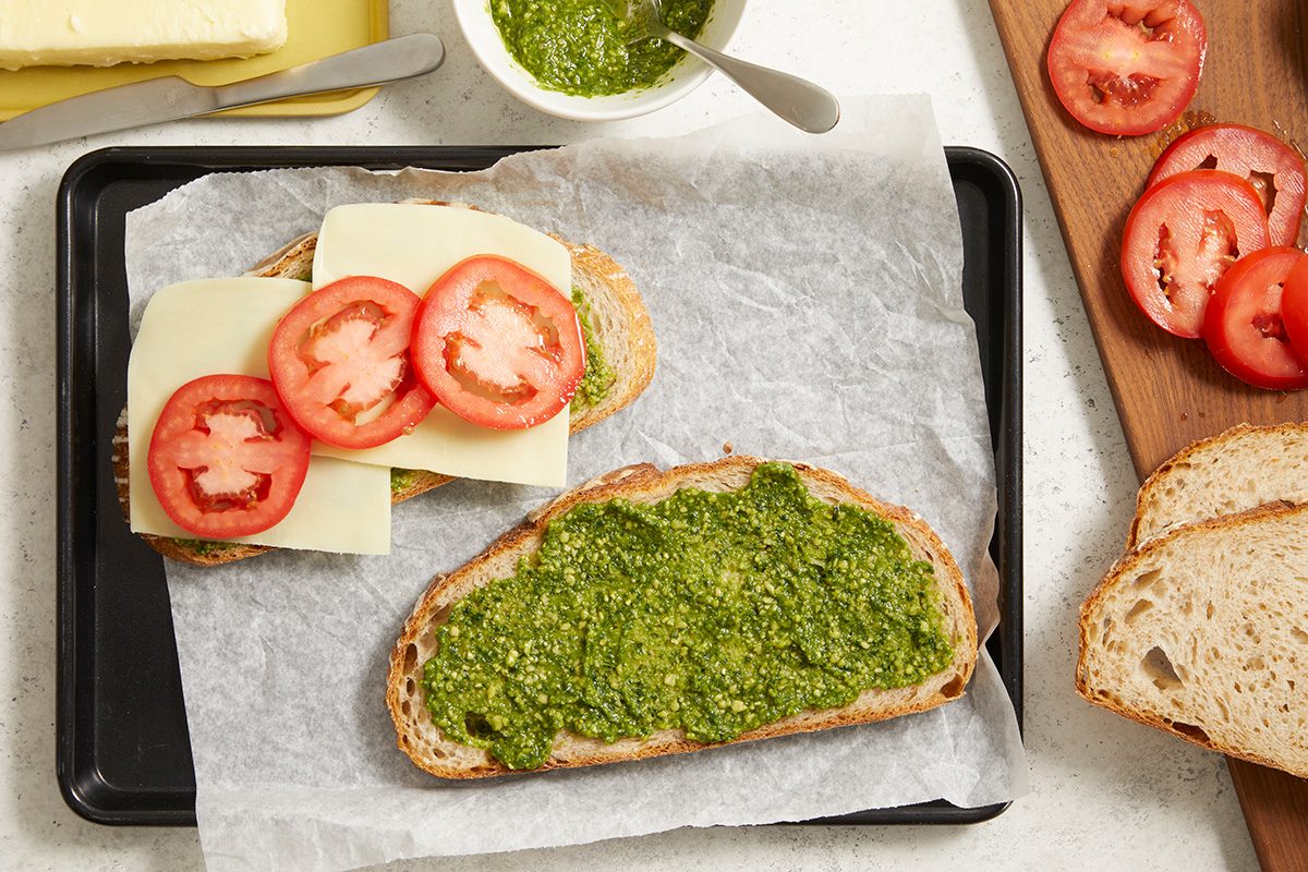 Slices of bread on parchment paper; one slice spread with green pesto, another topped with cheese and tomato slices. A bowl of pesto, butter, sliced tomatoes, and more bread are visible nearby.