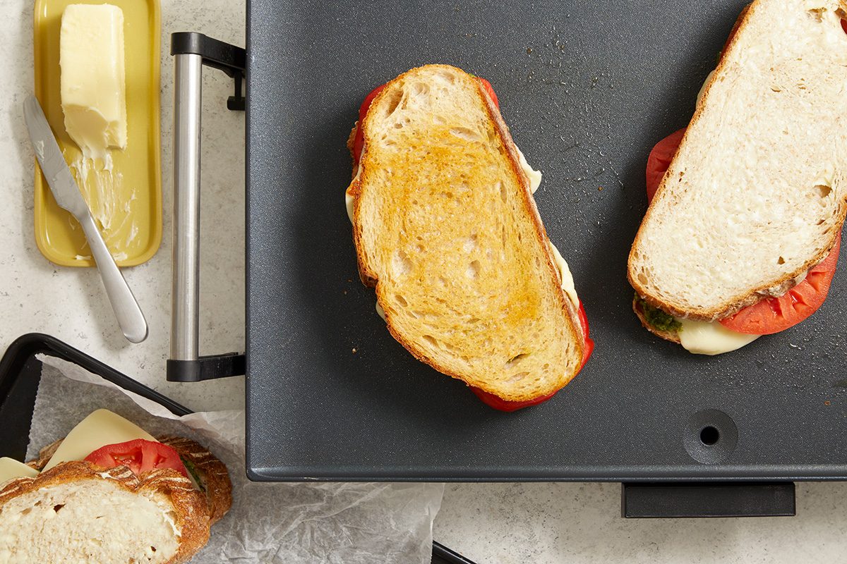 Two grilled cheese sandwiches with tomato are cooking on a griddle, with a butter dish and a butter knife nearby. Another sandwich is partially visible on a tray next to the griddle.